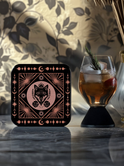 A set of four black, high-gloss square coasters featuring intricate rose-gold metallic illustrations of an owl, raven, stag, and wolf framed by celestial and botanical motifs. Made from durable masonite hardboard with cork backing for slip-resistant protection.